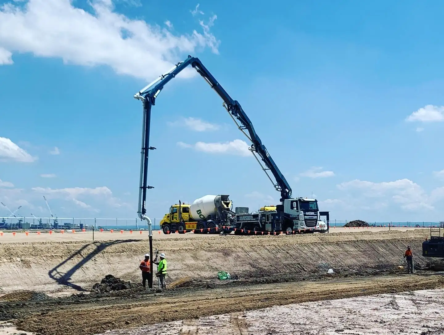 Worker guiding a boom pump hose to pour concrete footings, with a concrete truck feeding the pump