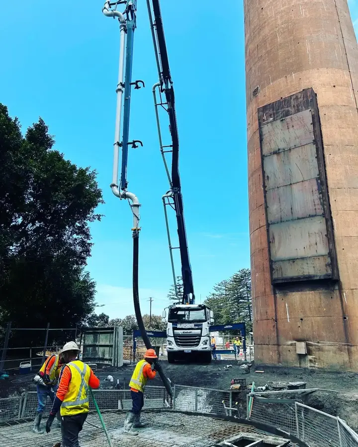 Crew placing concrete from a boom pump hose at the base of a tall brick chimney on a civil site